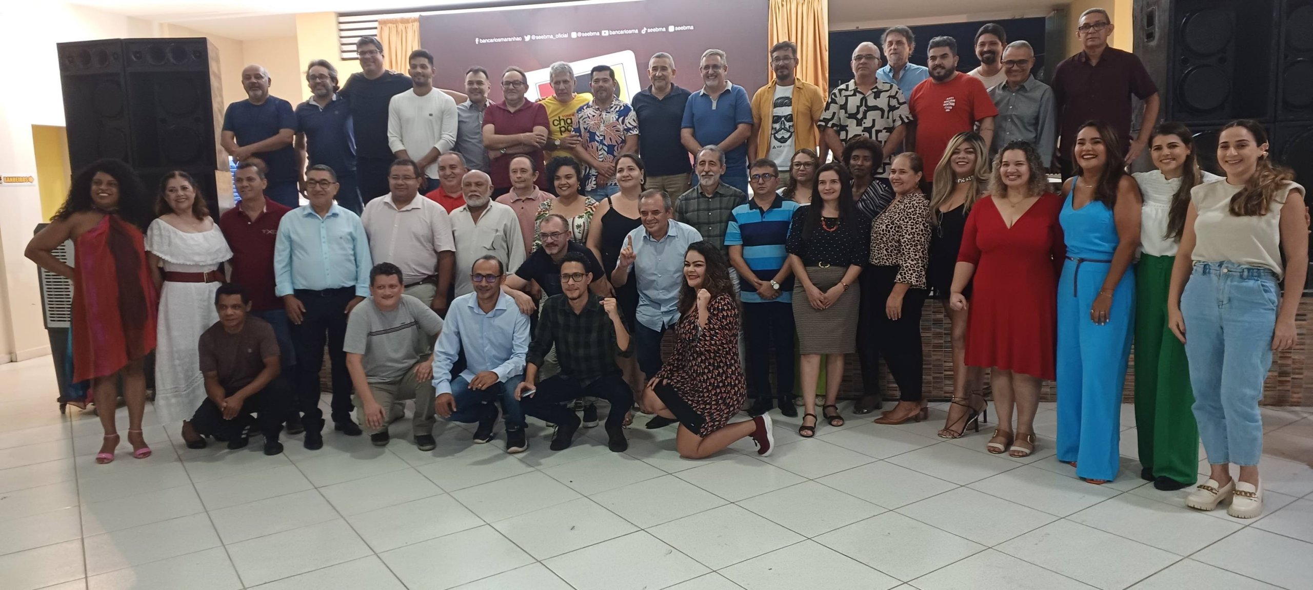 A imagem mostra a nova diretoria do Sindicato dos Bancários do Maranhão (SEEB-MA) reunida após a cerimônia de posse. O grupo é composto por diversas pessoas, tanto homens quanto mulheres, de diferentes idades. Eles estão dispostos em várias fileiras, com alguns agachados na frente e outros de pé atrás. A maioria dos presentes está sorrindo e vestida de maneira casual ou semi-formal. O fundo exibe um painel com o logotipo do sindicato e algumas caixas de som grandes estão visíveis nos lados esquerdo e direito da imagem. A atmosfera parece ser de celebração e camaradagem.